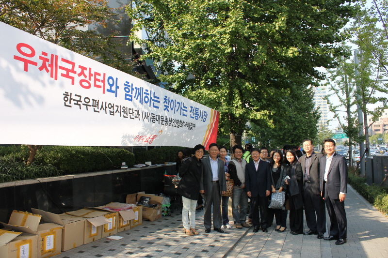 우체국장터와 함께 찾아가는 전통시장  / 한국우편사업지원단과 (사)동대문총상인연햡회 자매결연 / 우체국 쇼핑 / 행사 플랜카드 앞에 서있는 모습이다.