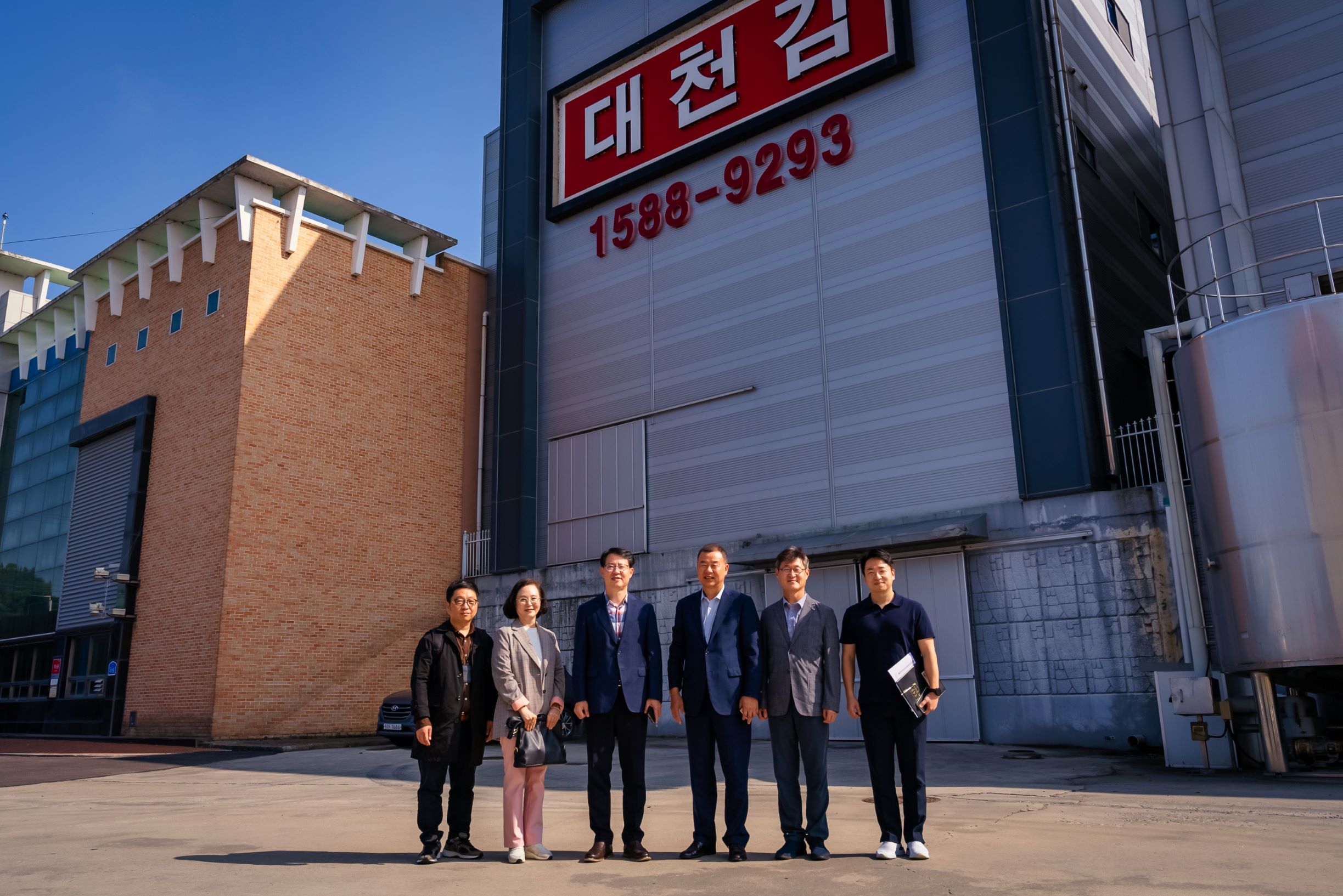 공급업체 건물 앞에 6명이 서서 기념촬영 중인 모습