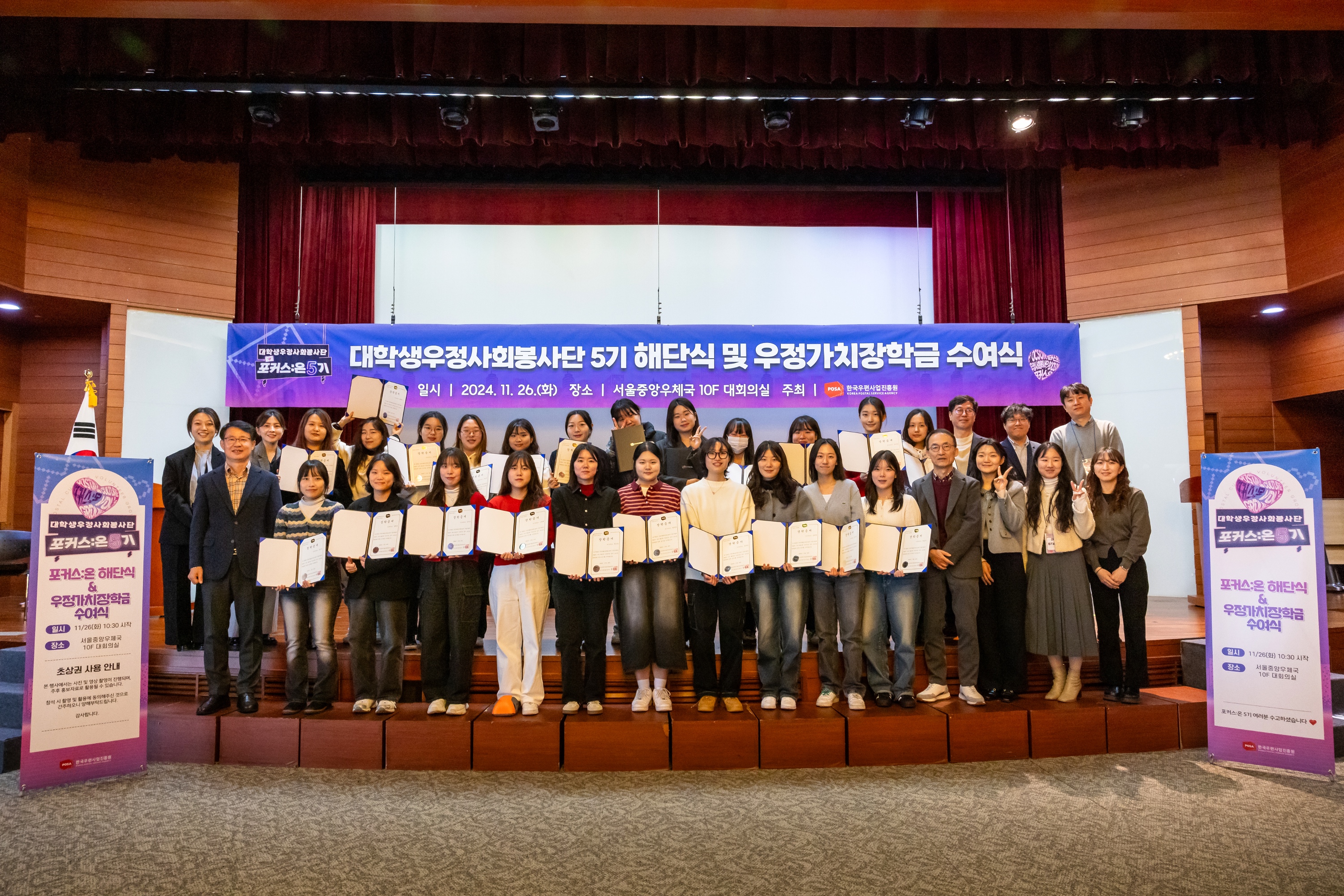 2024년 우정가치장학금 수상 상장을 펼쳐 강당 위에 두줄로 서서 기념촬영하는 모습