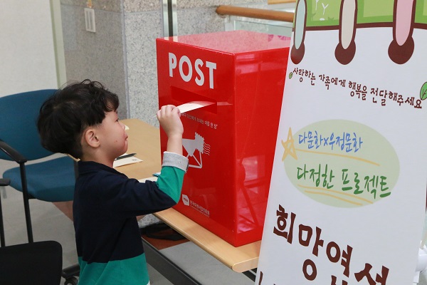 남자어린이가 쓴 편지를 우체통에 넣고 있다. 입간판 - 사랑하는 가족에게 행복을 전달해주세요,다문화X우정문화 다정한 프로젝트 희망엽서