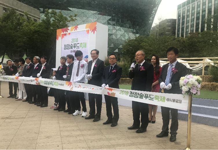 서울광장에서 열리는 '2018 청정숲푸드 축제 개막식 행사'에 참석하여 테이프 커팅식을 하고 있는 사람들과 마이크를 대고 말하고 있는 임정수 원장